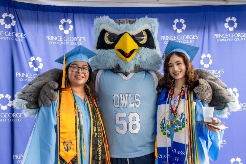 Two graduates in blue caps and gowns pose with an owl mascot at Prince George’s Community College commencement.