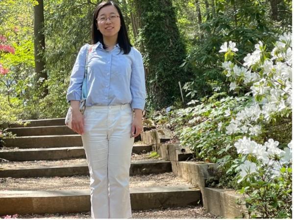 He Chen smiles at the camera in a sky blue button-up shirt and white pants in a sunny path.