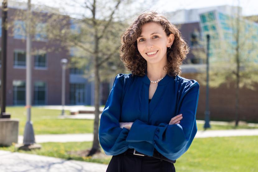 London Hudnall's curly hair glistens in the sunlight as she stands outside wearing a blue blouse with black pants. Behind her is the College's Bladen Hall building, several trees, and grassy areas.