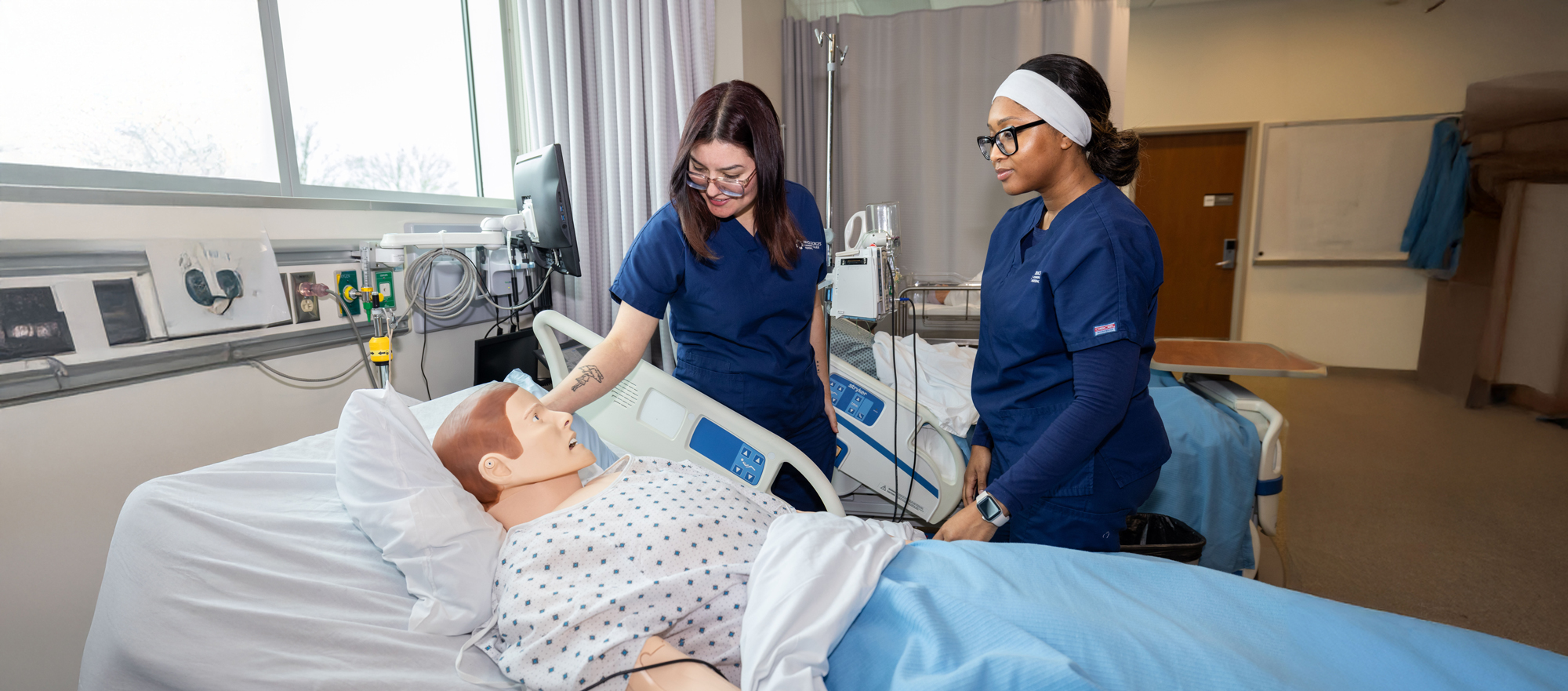 Gabriela Rivera and Radiatu Kamara in blue scrubs providing care to a patient simulation mannequin in a hospital training room.