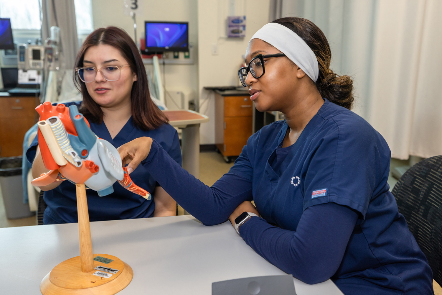 Two nursing students in blue scrubs studying a heart model together in a clinical skills lab.