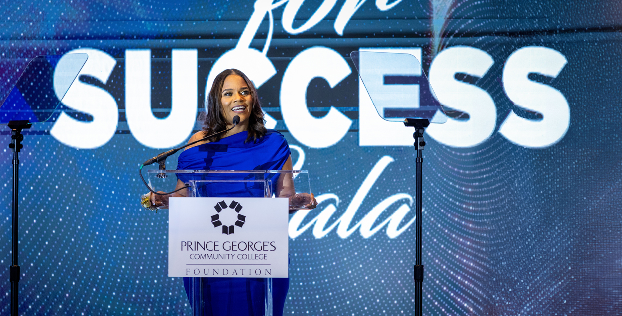 Dena Wilson speaks at a clear podium labeled “Prince George’s Community College Foundation,” with an illuminated “Partners for Success Gala” backdrop behind her.