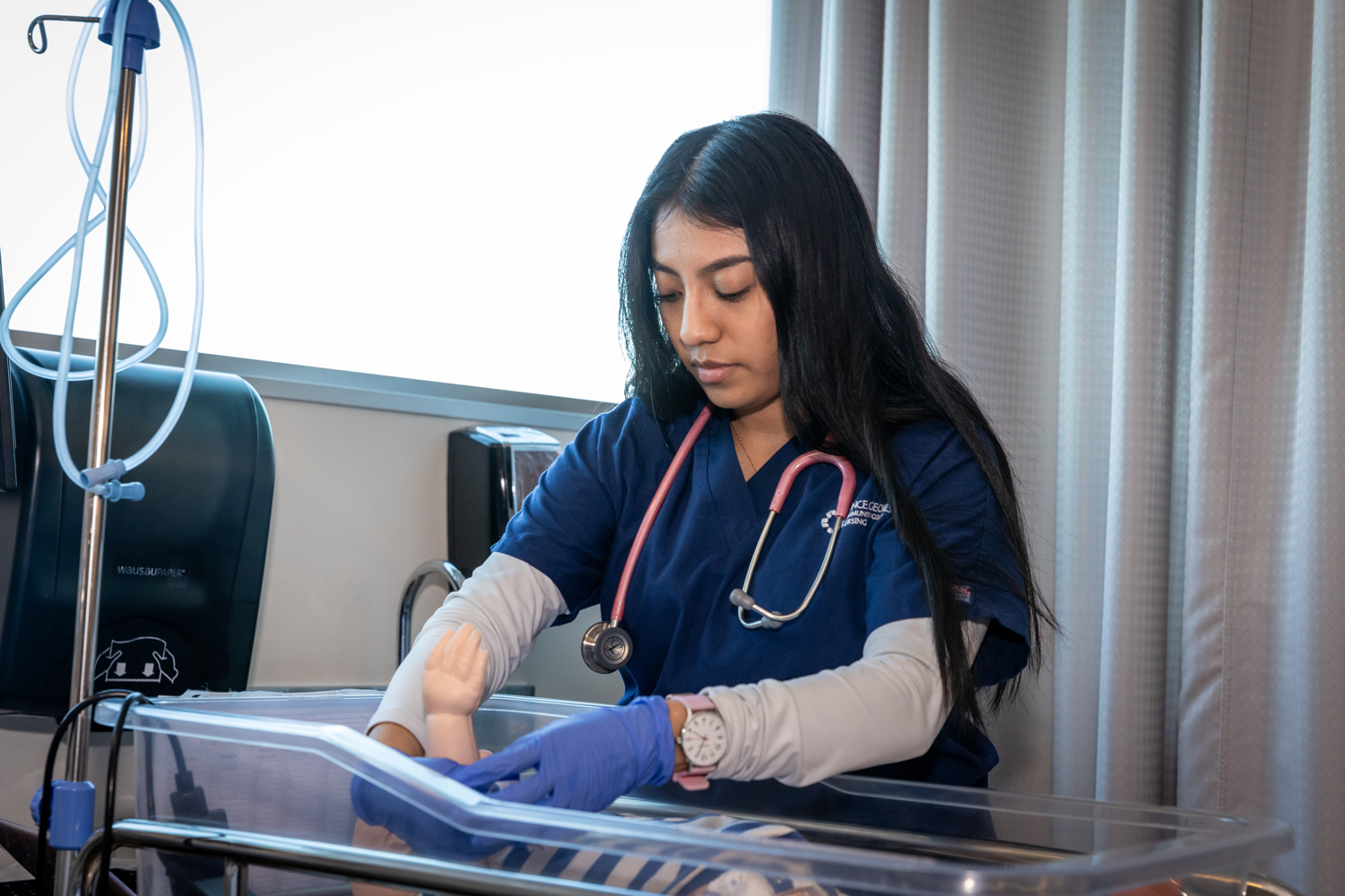 Nursing student in blue scrubs practicing newborn care on a simulation mannequin in a lab setting.