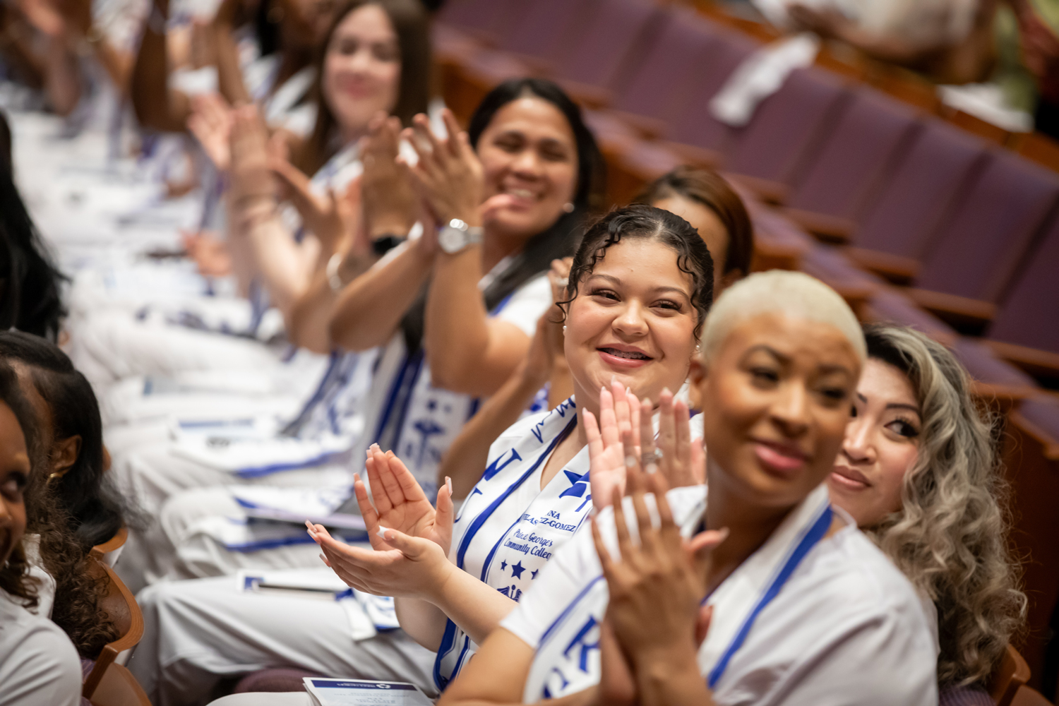 Nursing graduates in white uniforms and blue stoles clapping and smiling during a ceremony.