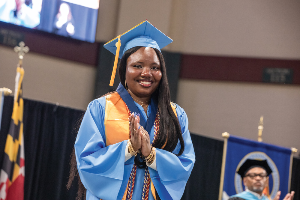 Ahnora smiles and clasps her hands while wearing a blue cap and gown with cords and a gold stole during 2025 commencement.