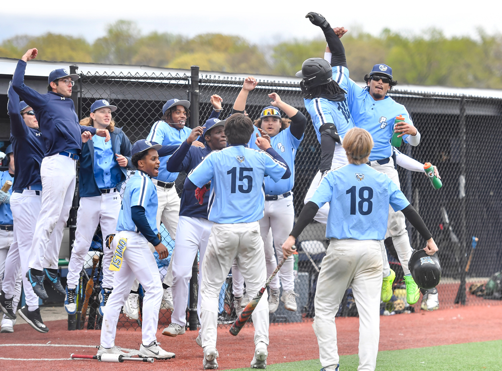 PGCC baseball players in light blue jerseys celebrate excitedly near the dugout after a home run, jumping and raising their arms as teammates gather around home plate.