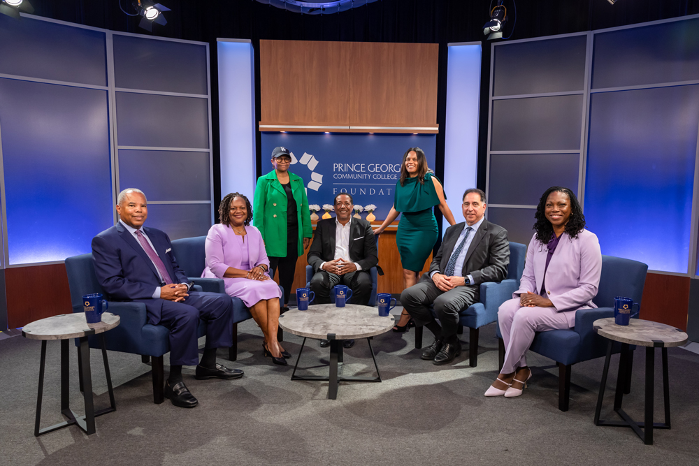 Foundation Board Members in a studio set with a “Prince George’s Community College Foundation” backdrop, arranged in a semicircle of blue chairs around small tables with branded mugs.