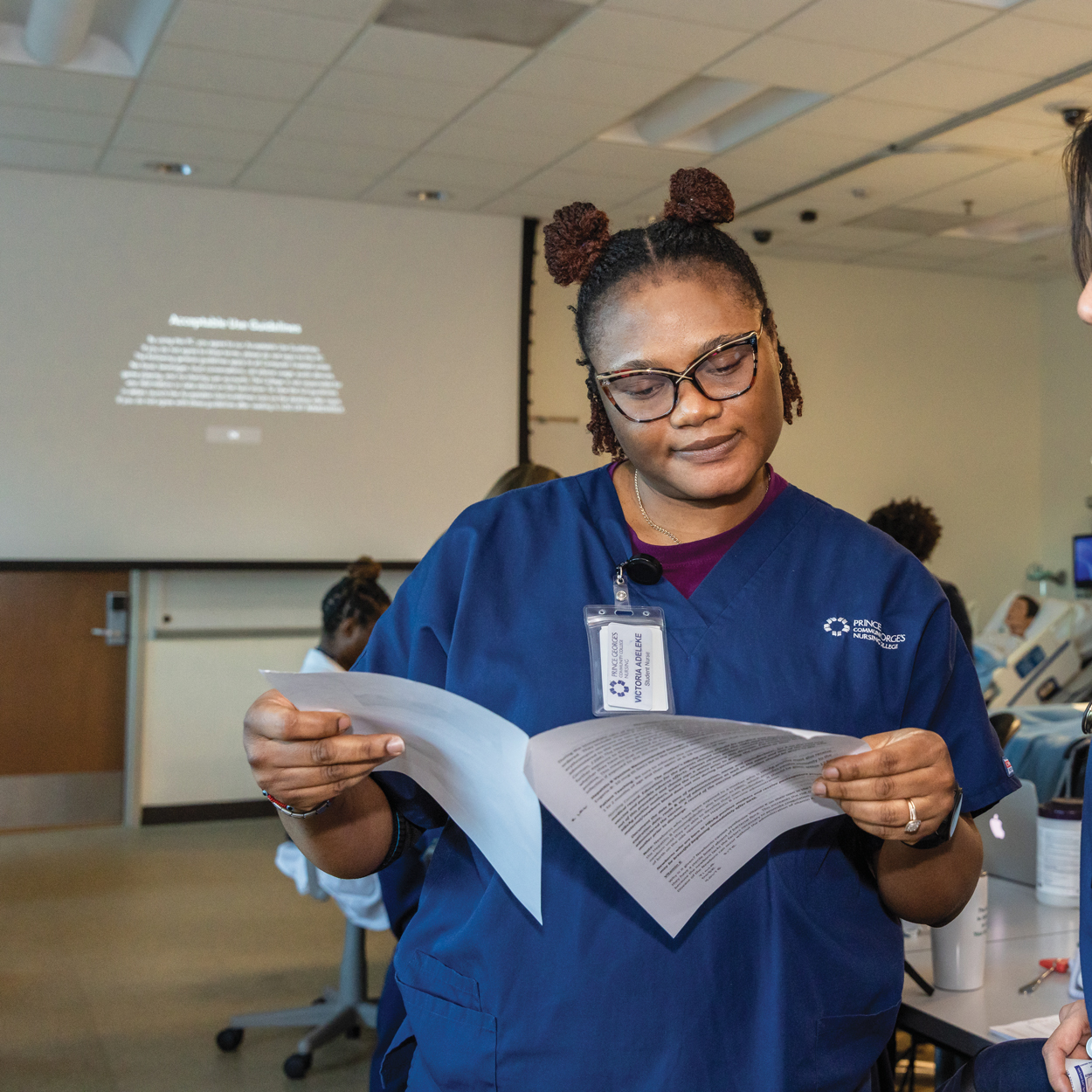 Victoria Adeleke blue nursing scrubs reviewing papers in a classroom clinical training setting.