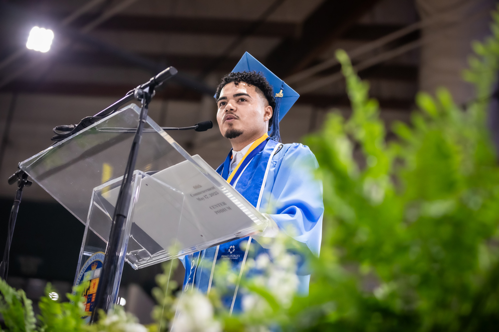 William Flores, 2025 Distinguished Graduate, speaks at a clear podium wearing a blue cap and gown with honor cords, delivering a speech at a commencement ceremony.