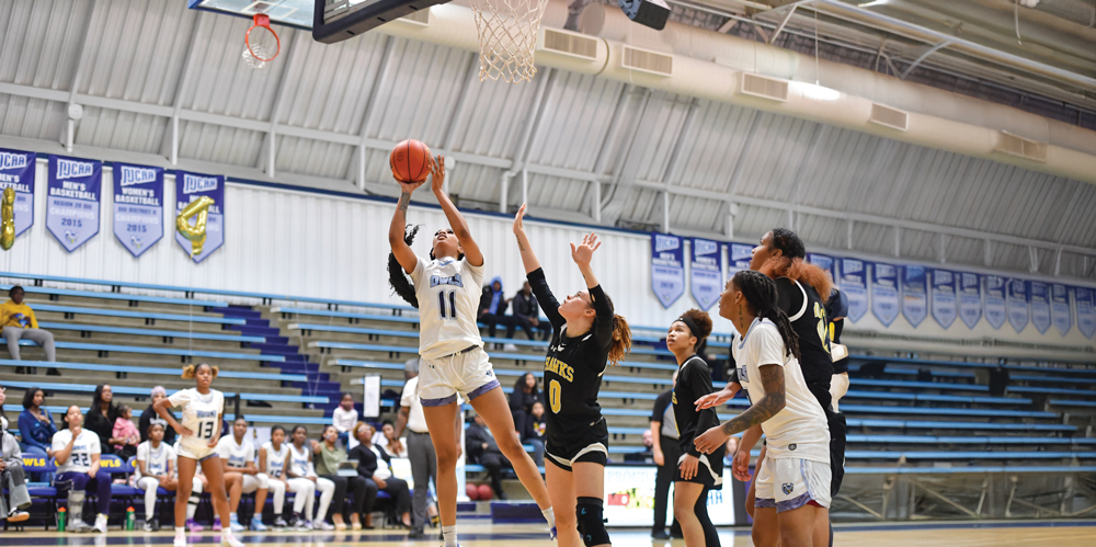 A women’s basketball player in a white uniform leaps toward the basket for a layup while defenders in black uniforms reach up to defend the shot during a game in a gymnasium.