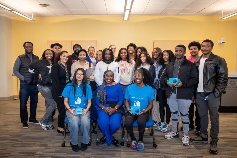 Group of students and instructors posing together in a classroom during a cybersecurity training event, with several participants holding small tech devices.