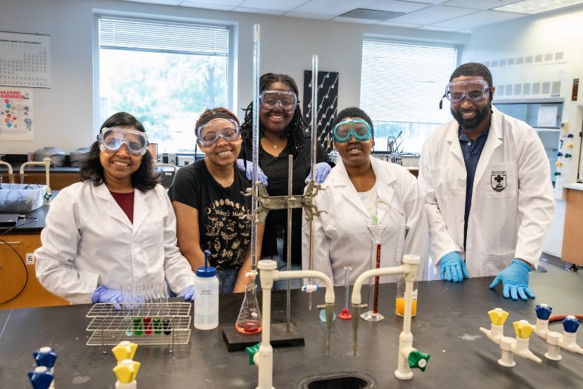 Dr. Sumana Yasapala and students wearing lab coats and safety goggles, standing together in a chemistry lab with glassware and equipment on the table.