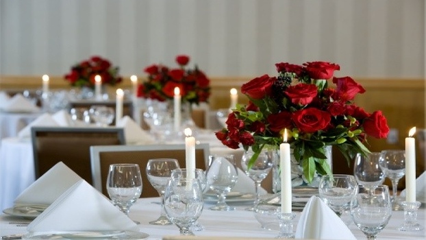 Elegant banquet tables set with white linens, red rose centerpieces, glassware, and lit candles in a formal dining setting. For the use of hospitality