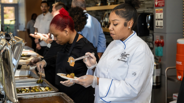 Culinary students serving food from buffet trays in a kitchen setting.