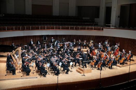 The PGCC Philharmonic performs onstage in a large concert hall. Dozens of musicians in formal black attire play strings, woodwinds, brass, percussion, and harp, with a conductor leading from a podium. Warm lighting and a festive holiday atmosphere fill the space.